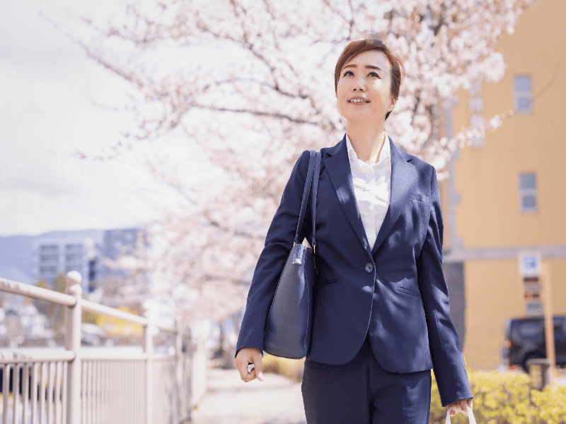 桜の木を背景に新人女性行政書士がさわやかな笑顔で歩いている写真